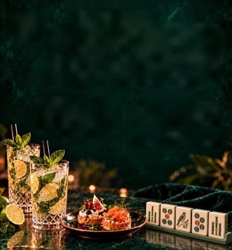 Mojitos with fresh mint, small bites on a copper plate and a row of mahjong tiles on a dark green marble table.